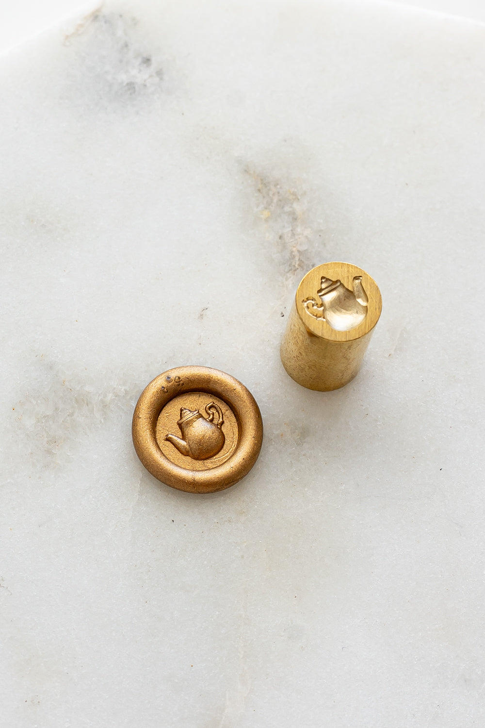 A mini brass wax seal stamp head with a 3D tea pot design beside a gold wax seal impression on a white marble surface