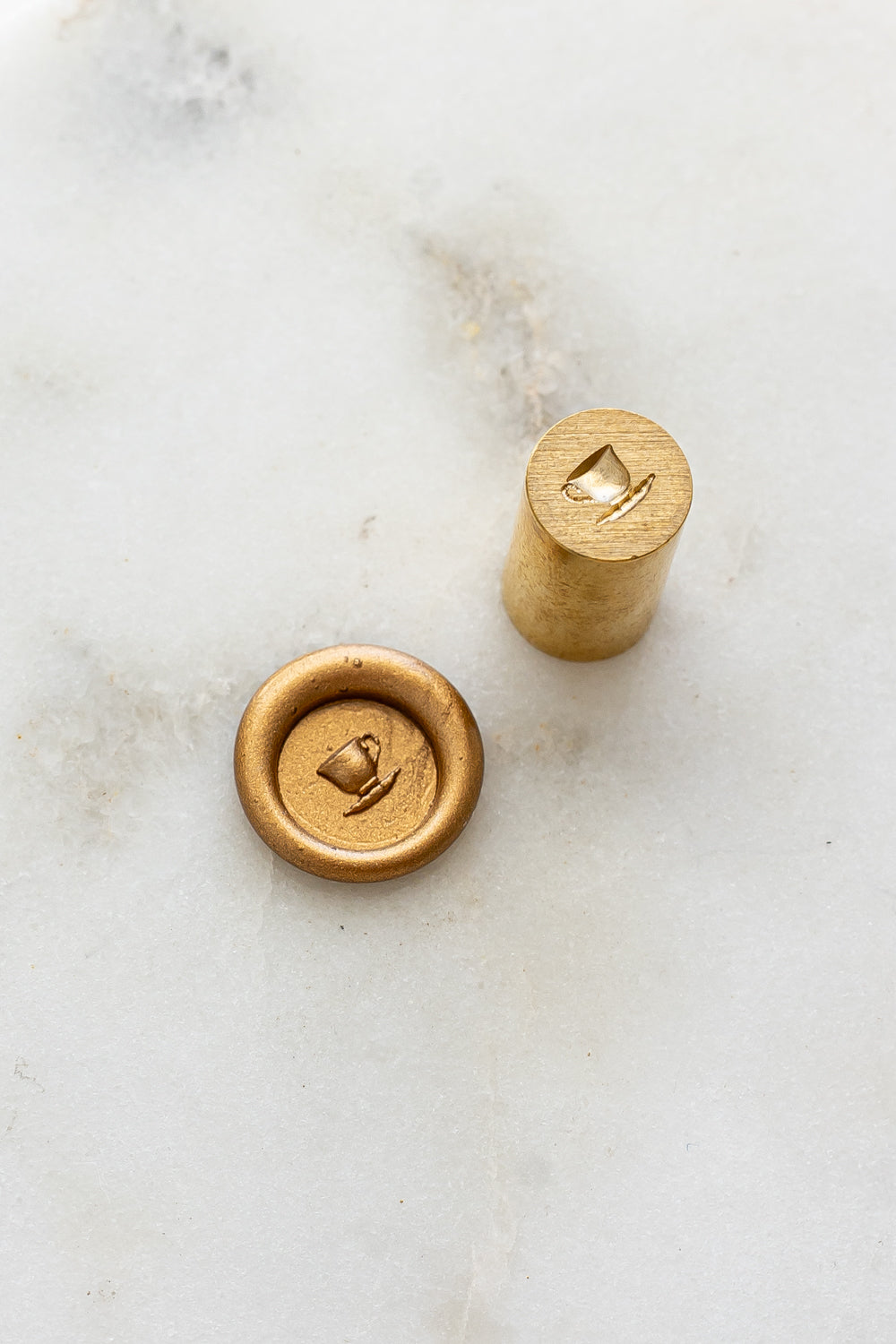 ​A mini brass wax seal stamp head with a 3D tea cup design beside a gold wax seal impression on a white marble surface