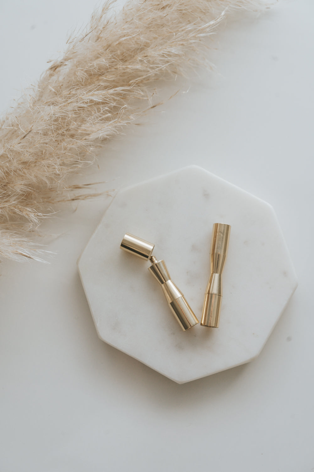 Two gold mini wax seals with removable heads on a white marble surface with dried pampas grass.