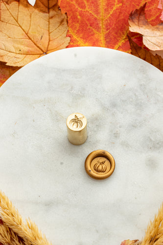 Gold mini wax seal stamp engraved with a pumpkin, shown beside its wax impression on a marble surface surrounded by fall leaves and wheat stems.