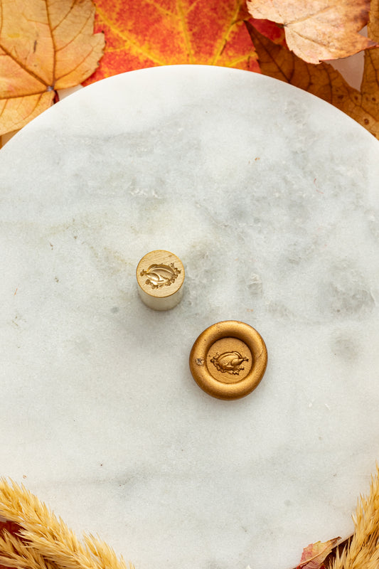 Gold mini wax seal stamp engraved with a Thanksgiving turkey, shown beside its wax impression on a marble surface surrounded by warm fall leaves and wheat stems.