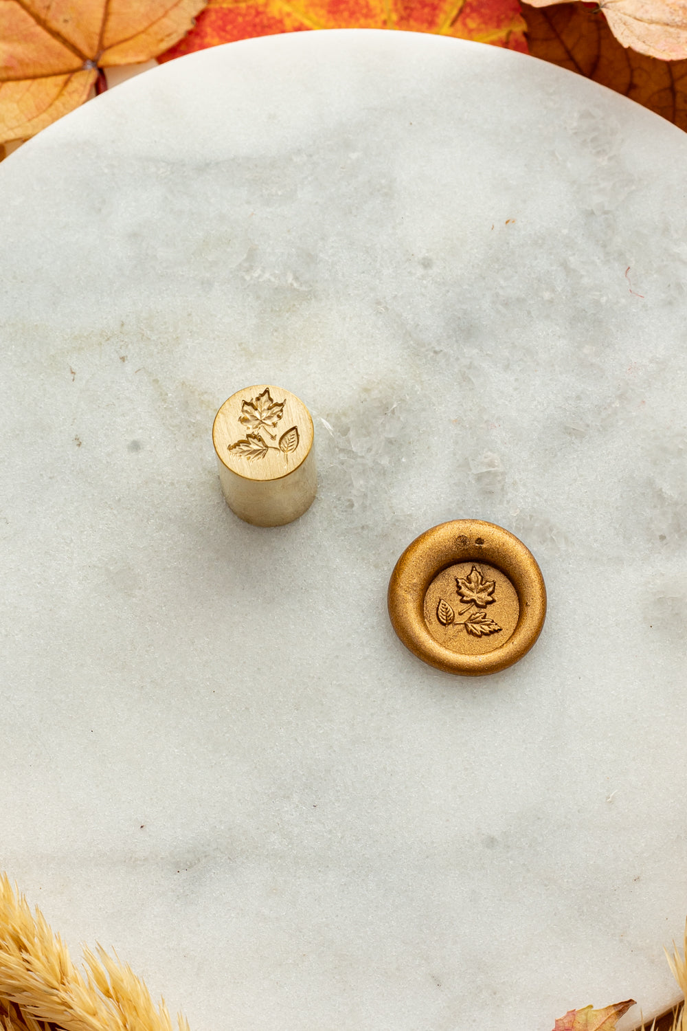 Gold mini wax seal stamp engraved with autumn leaves, shown beside its wax impression on a marble surface surrounded by golden wheat stems and colorful fall foliage.