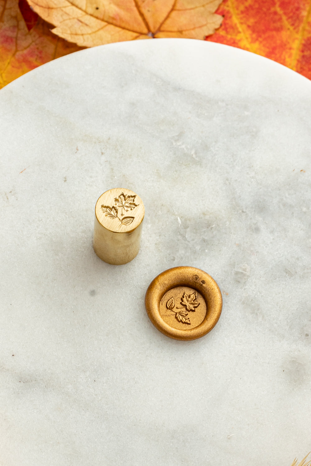 Gold mini wax seal stamp engraved with autumn leaves, shown beside its wax impression on a marble surface surrounded by golden wheat stems and colorful fall foliage.