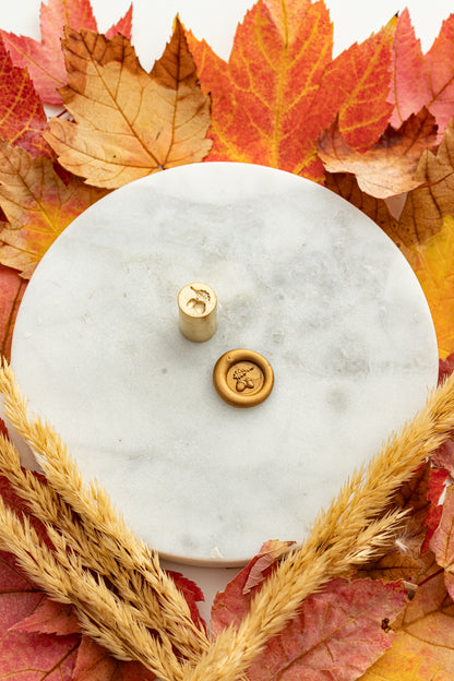 Gold mini wax seal stamp engraved with an acorn, shown beside its wax impression on a marble surface surrounded by autumn leaves and golden wheat stems.