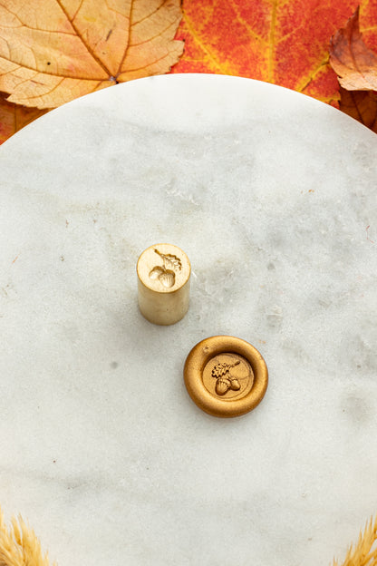 Gold mini wax seal stamp engraved with an acorn, shown beside its wax impression on a marble surface surrounded by autumn leaves and golden wheat stems.
