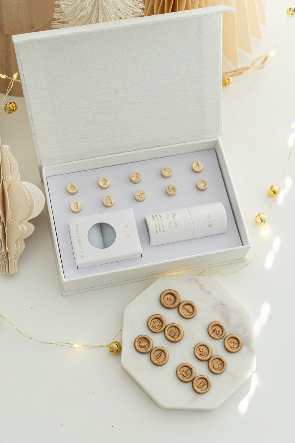Holiday Mini Wax Seal Head Set opened to reveal twelve brass stamp designs, paired with their gold wax seal impressions on a marble slab, surrounded by soft festive décor and string lights.