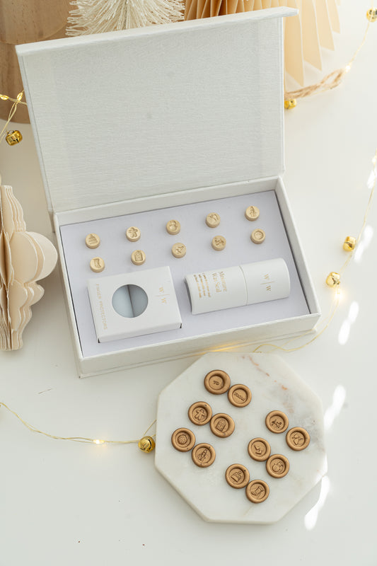 Holiday Mini Wax Seal Head Set opened to reveal twelve brass stamp designs, paired with their gold wax seal impressions on a marble slab, surrounded by soft festive décor and string lights.