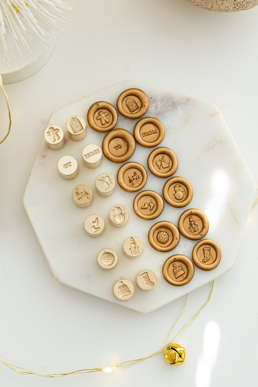Flat lay of the Holiday Mini Wax Seal Head Set showing all twelve festive brass stamp designs and their matching wax seal impressions on a marble slab with gold holiday lights.