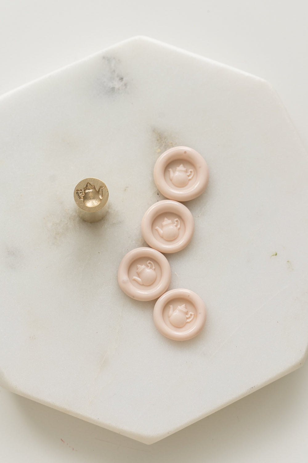 A mini brass wax seal stamp head with a 3D tea pot design beside a bunch of blush mini wax seals on a white marble coaster
