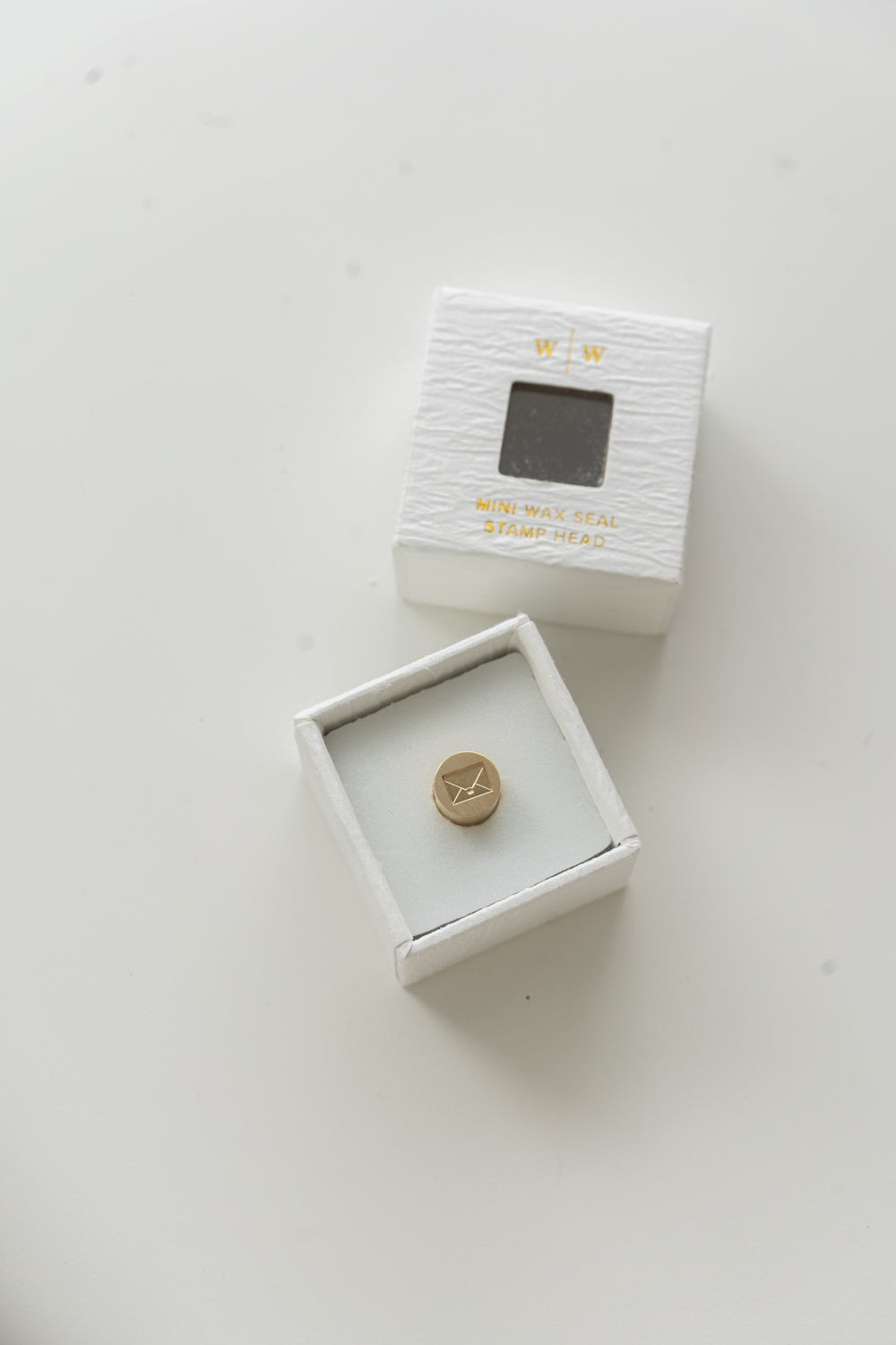 ​A mini brass wax seal stamp head with an envelope design in a textured white ring-style box, with its lid beside it