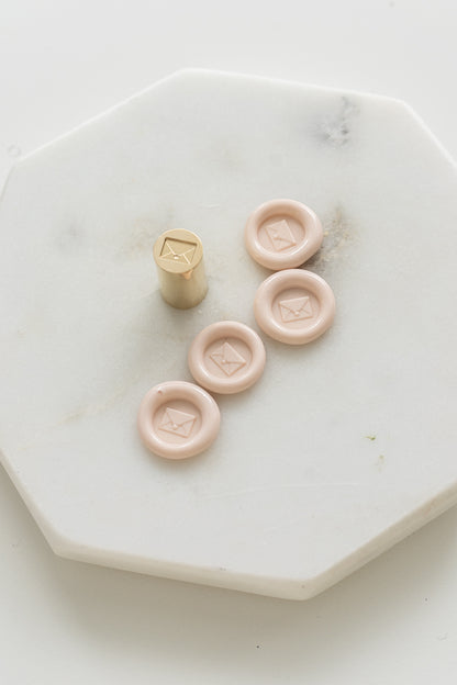​​A mini brass wax seal stamp head with an envelope design beside a bunch of blush mini wax seals on a white marble coaster