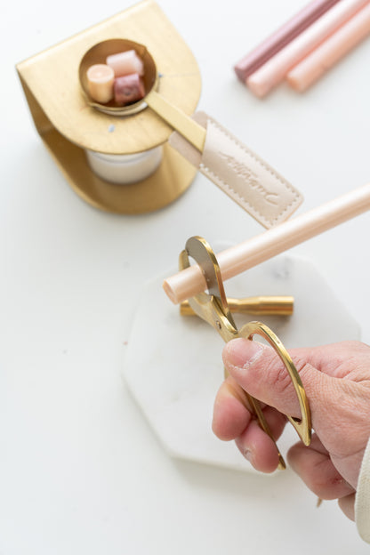 Hand using a gold sealing wax trimmer on a white surface with a brass wax seal stove