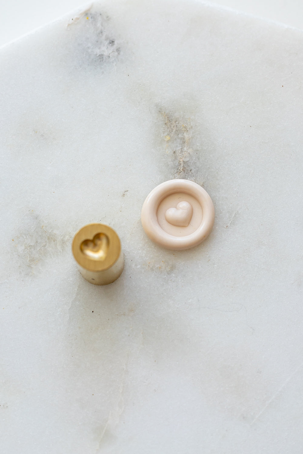A mini brass wax seal stamp head with a 3D heart design beside a blush wax seal impression on a white marble surface