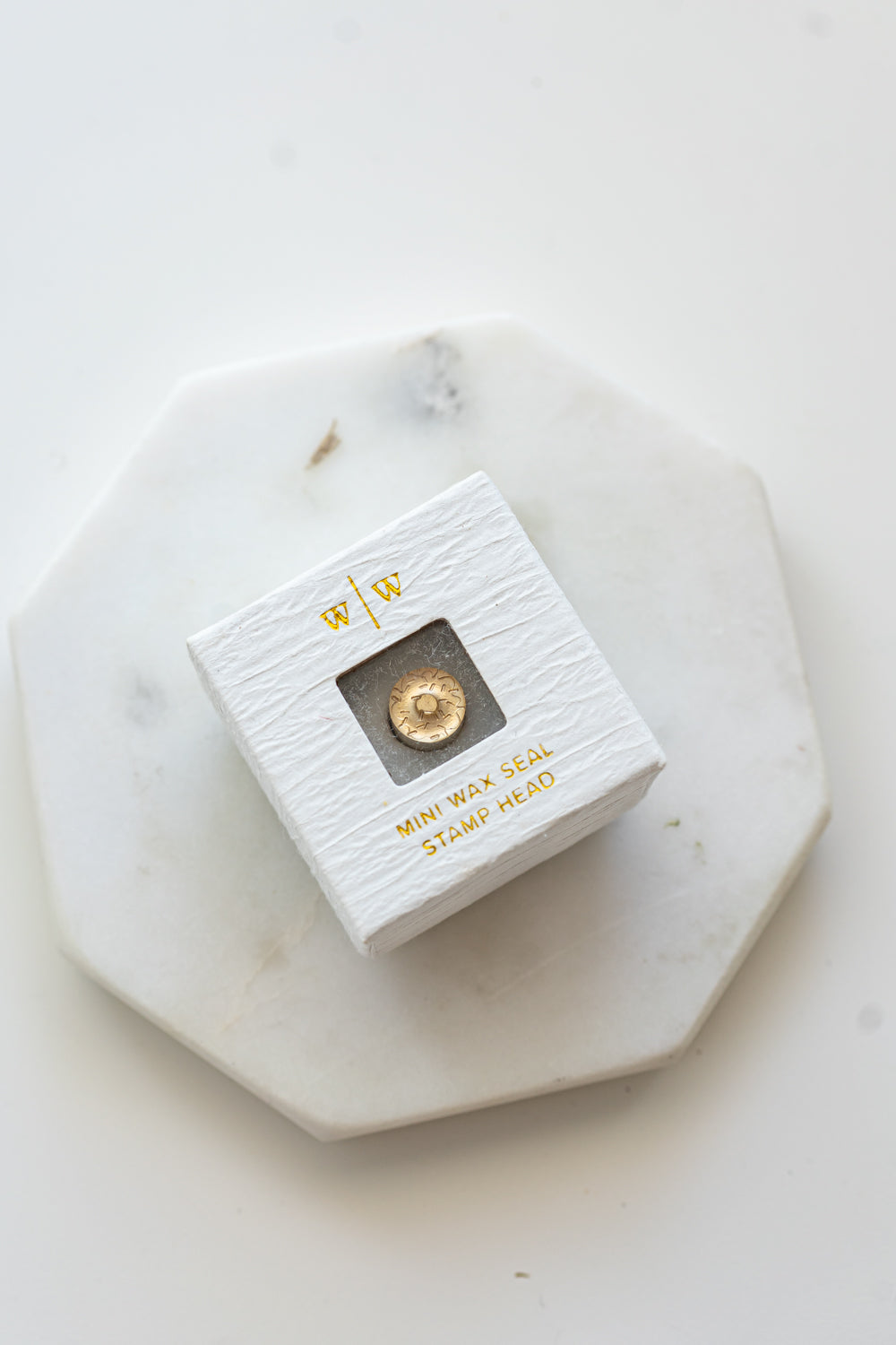 A mini brass wax seal stamp head with a 3D donut design in a textured white ring-style box on a marble coaster