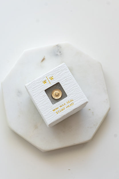 A mini brass wax seal stamp head with a 3D donut design in a textured white ring-style box on a marble coaster