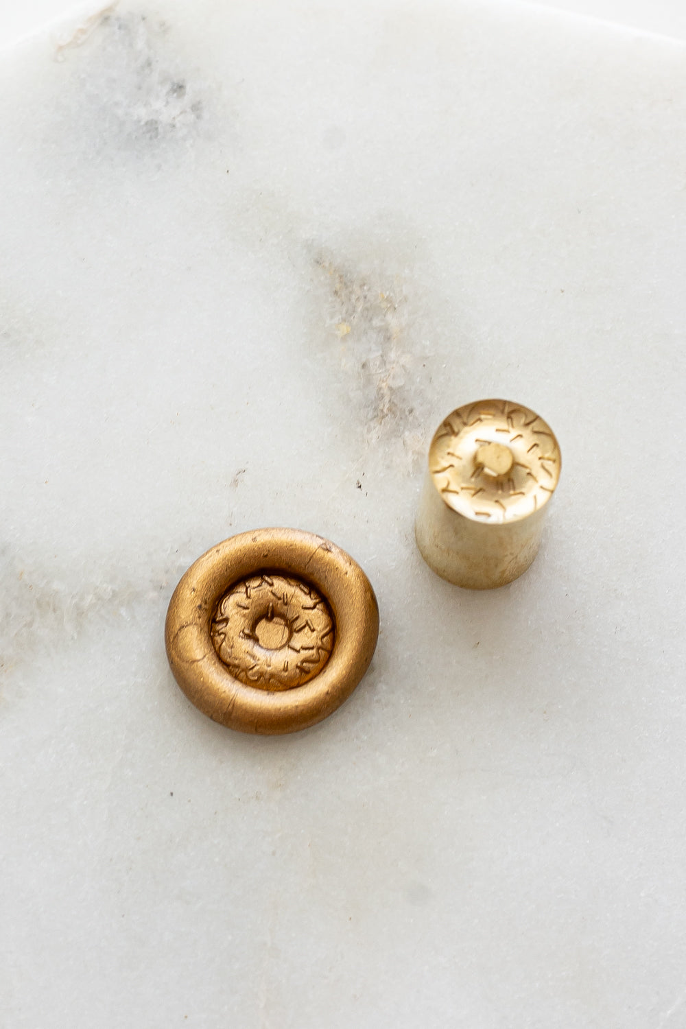 ​A mini brass wax seal stamp head with a 3D donut design beside a gold wax seal impression on a white marble surface