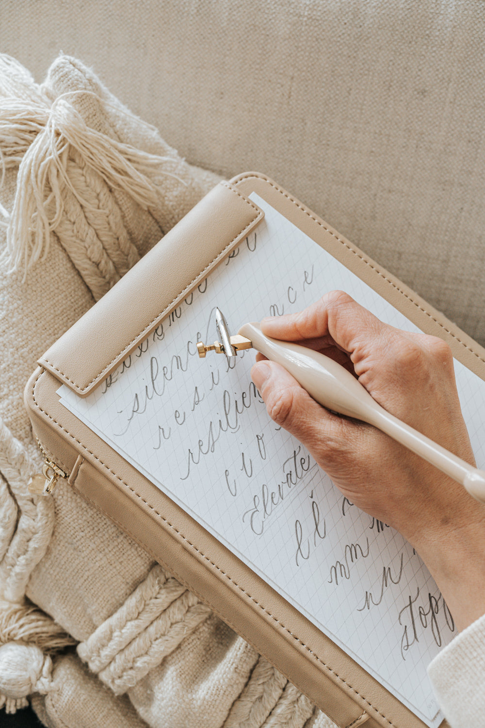 A hand practicing calligraphy with an ivory oblique pen on a gridded sheet, resting on a beige Calligraphy Travel Kit case.