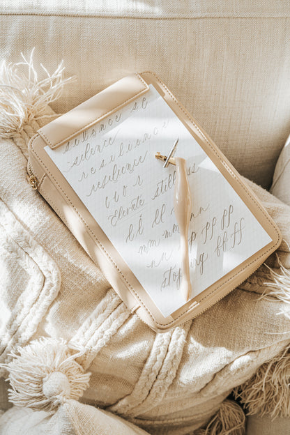 Beige Calligraphy Travel Kit with a practice sheet and blush oblique pen, resting on a textured neutral cushion.