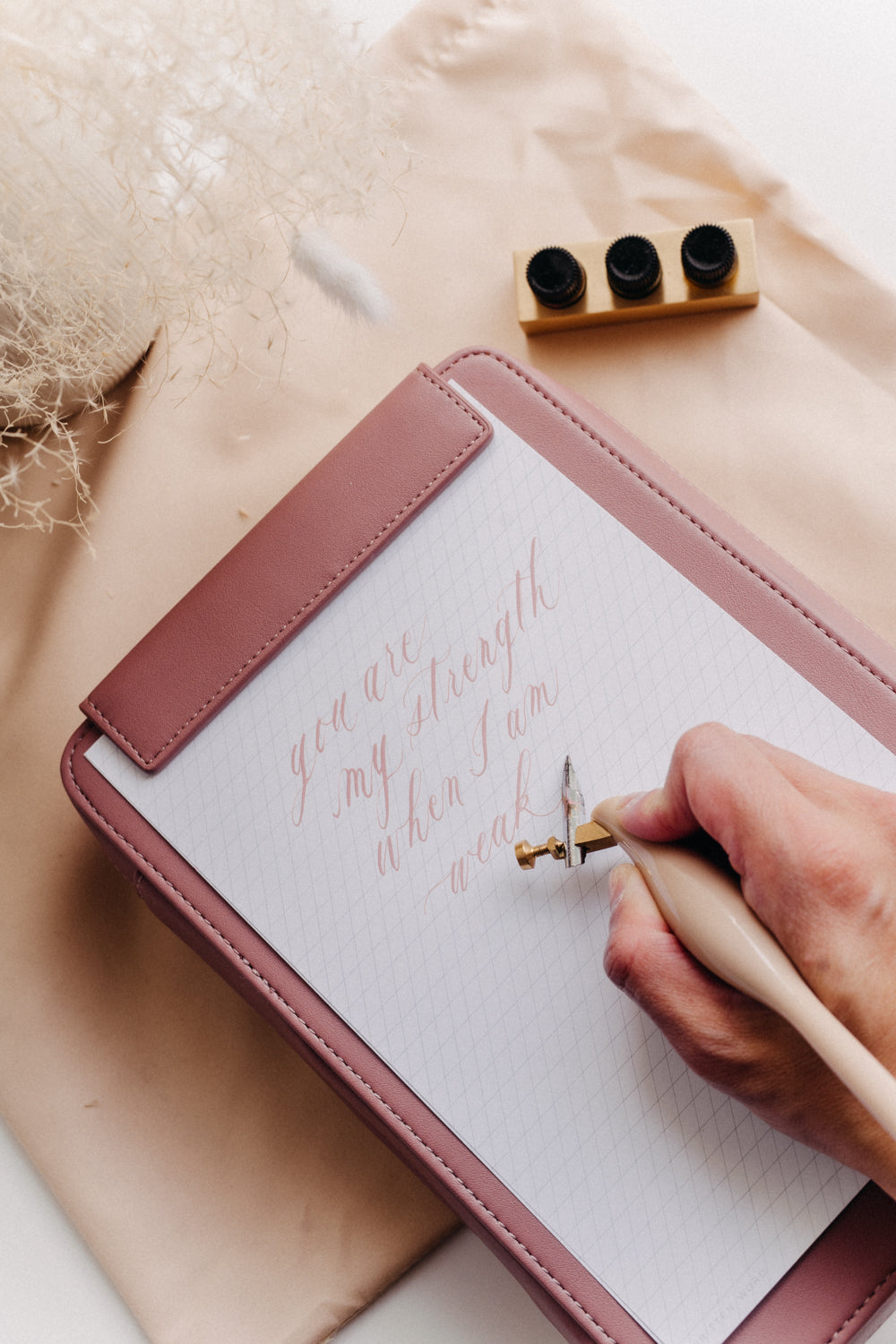 A dusty rose travel kit on a beige dust bag with a slanted guide notepad and mini tilted inkwell on a white surface