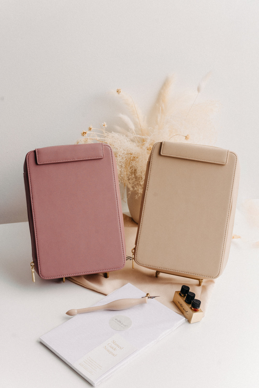 A dusty rose and beige travel calligraphy case on a white table along with a notepad, oblique pen, inkwell, and dried florals
