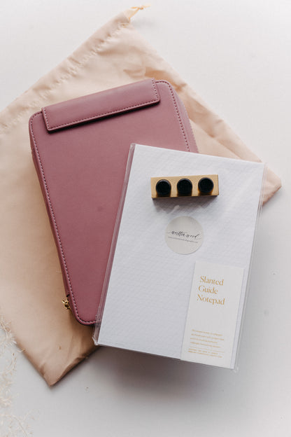 A dusty rose travel kit on a beige dust bag with a slanted guide notepad and mini tilted inkwell on a white surface