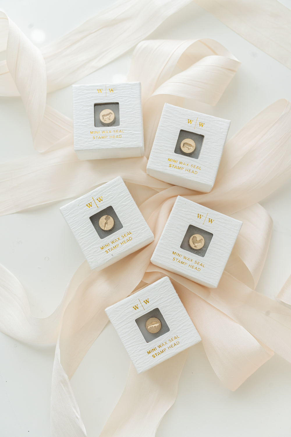 5 white ring-style boxes with mini wax seal heads featuring a cow, pig, carrot, chicken, and fish designs, neatly arranged on top of a blush ribbon on a white surface