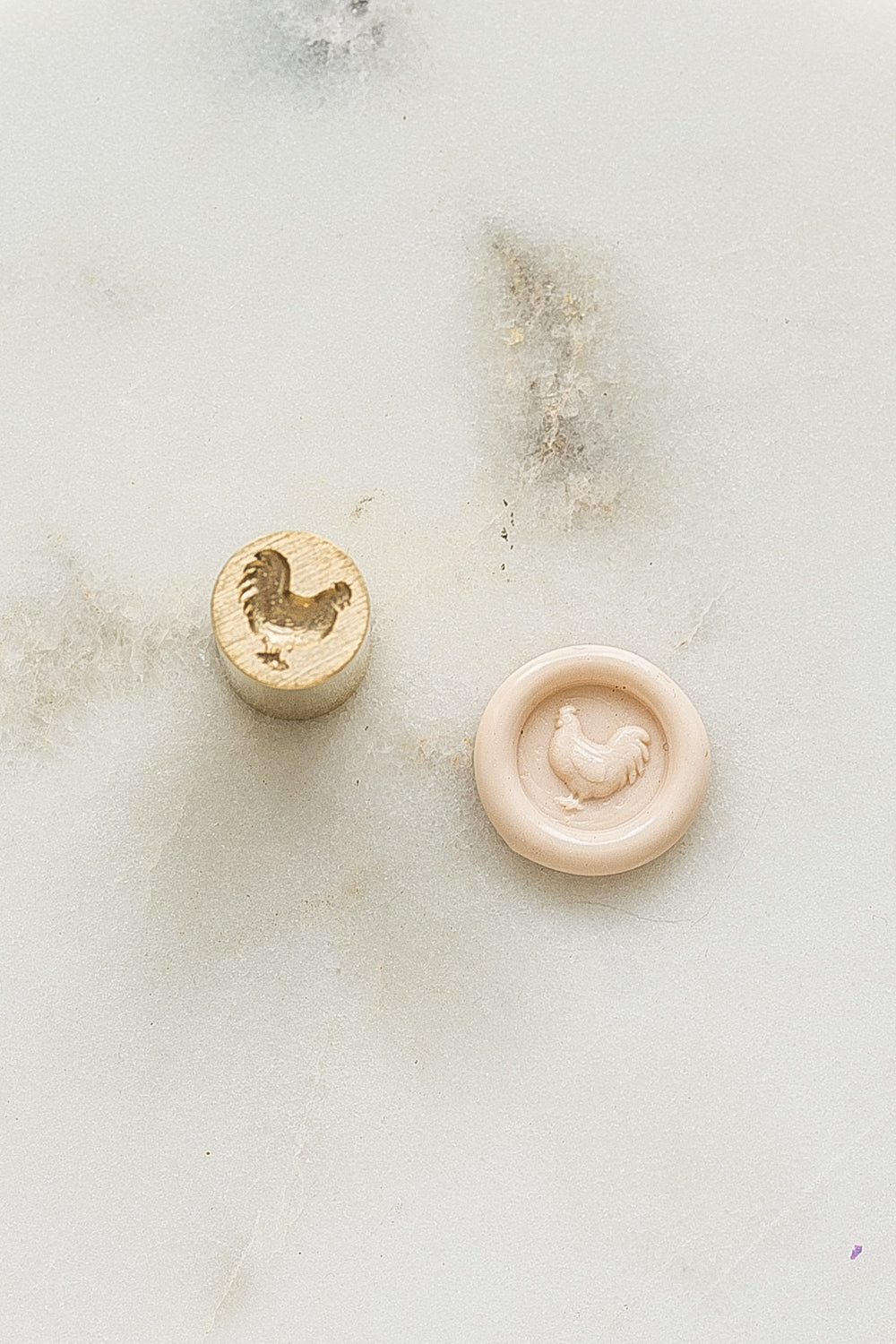 ​A mini brass wax seal stamp head with a 3D chicken design beside a blush wax seal impression on a white marble surface