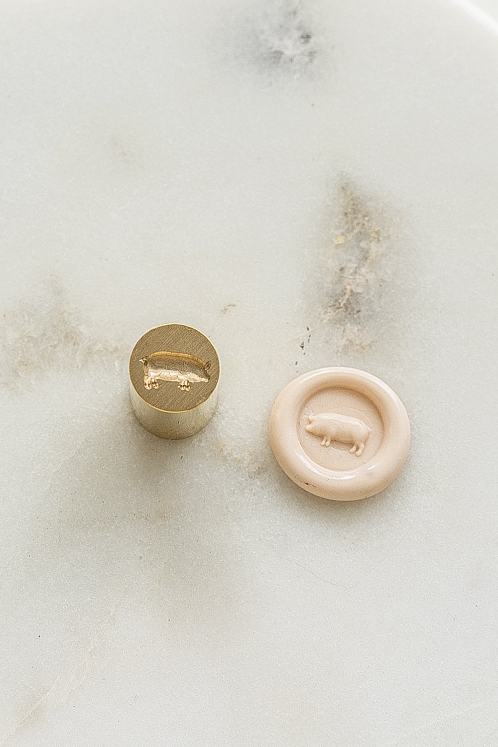 ​A mini brass wax seal stamp head with a 3D pig design beside a blush wax seal impression on a white marble surface
