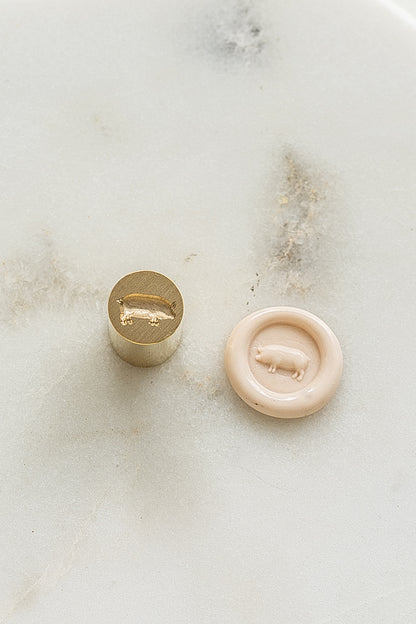 ​A mini brass wax seal stamp head with a 3D pig design beside a blush wax seal impression on a white marble surface