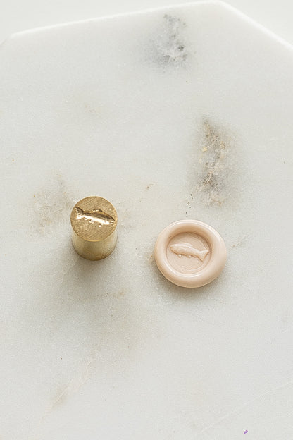 A mini brass wax seal stamp head with a 3D fish design beside a blush wax seal impression on a white marble surface
