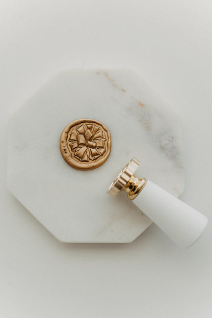 Gold gift bow wax seal on white marble tray beside brass and wooden wax seal stamp, top view