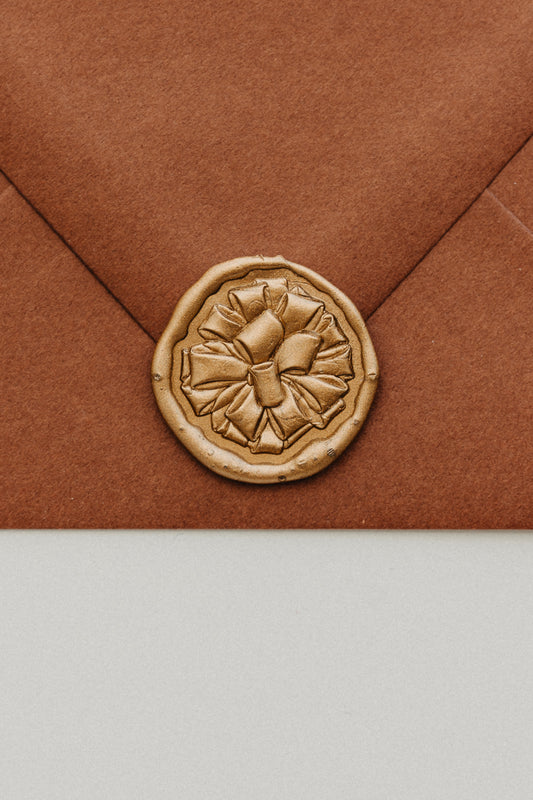 Close-up of gold gift bow wax seal on rust terracotta envelope flap, holiday card sealing