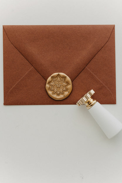 Gold geometric star wax seal securing a warm brown envelope beside a white-handled brass stamp.