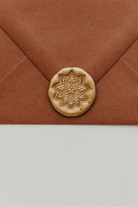 Close-up of a gold geometric star wax seal securing the flap of a warm brown envelope.