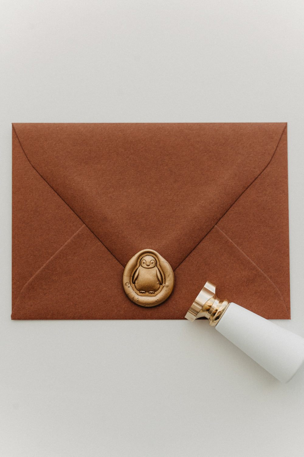 Gold penguin wax seal on a warm brown envelope beside a white-handled brass wax seal stamp.
