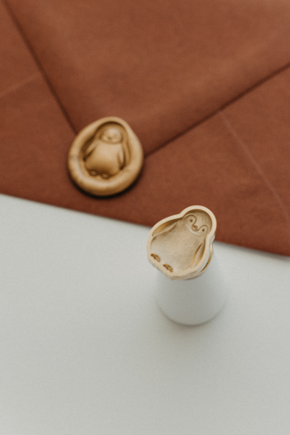 White-handled brass penguin wax seal stamp displayed beside a gold wax-sealed warm brown envelope.