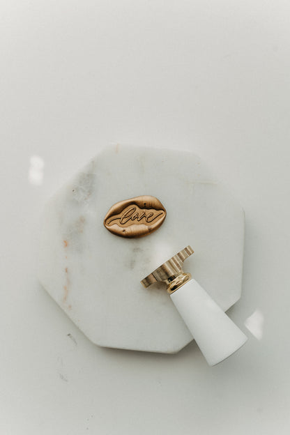 Gold “love” wax seal and white-handled brass wax seal stamp on a white marble coaster.