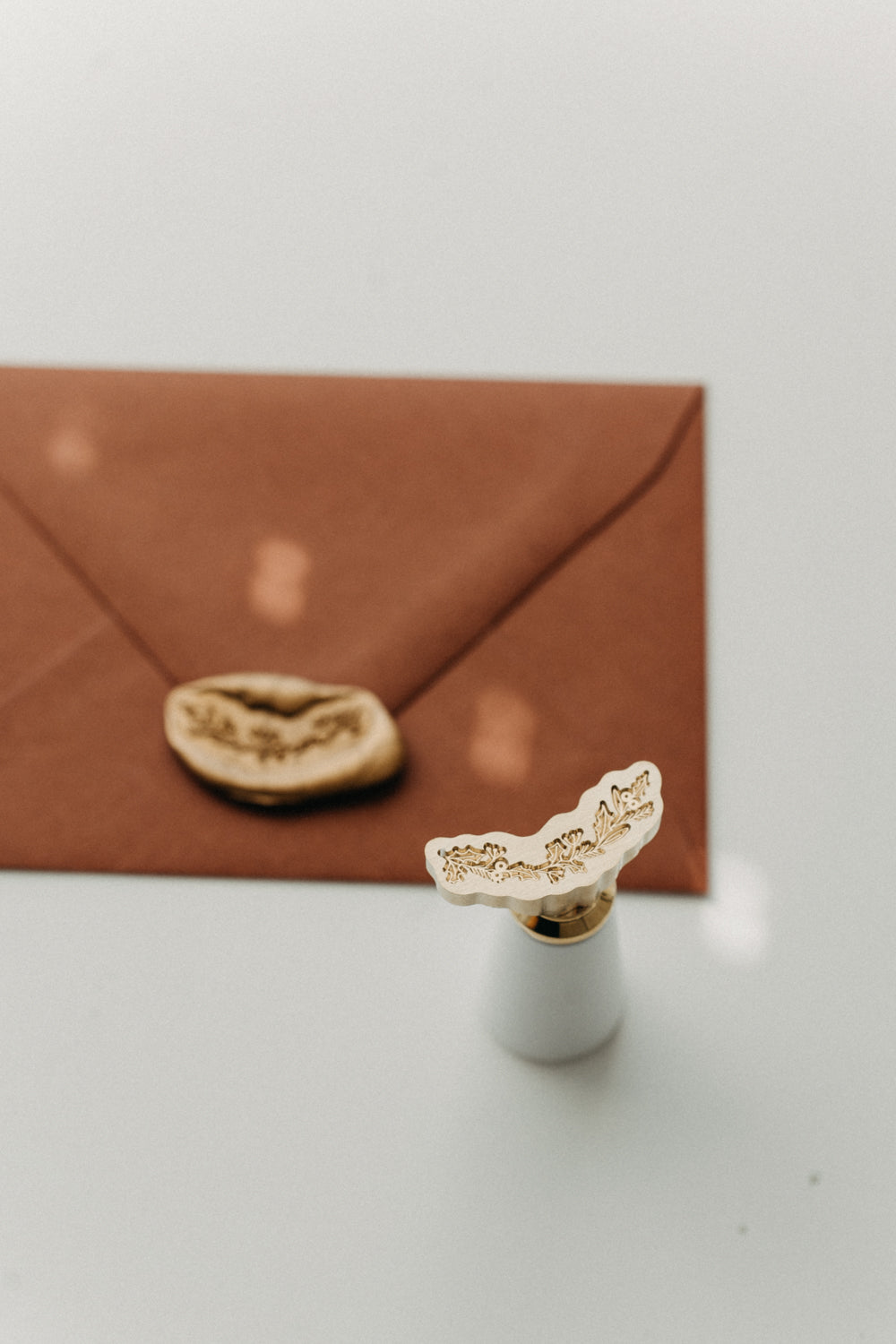 White-handled brass botanical garland wax seal stamp beside a gold wax-sealed warm brown envelope.
