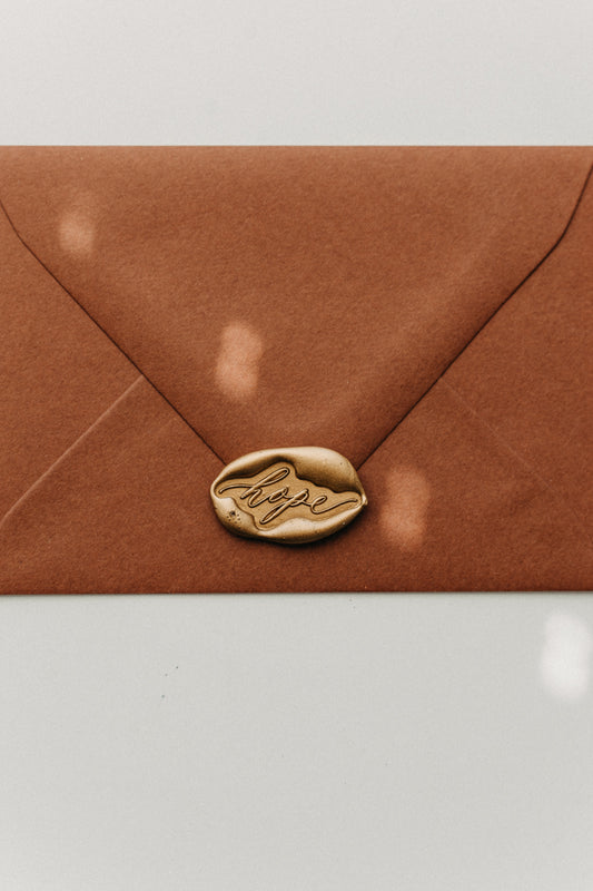 Metallic gold “hope” wax seal securing a warm brown envelope, close-up stationery detail.