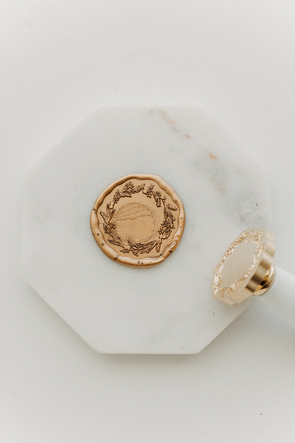 Metallic gold wax seal embossed with a botanical wreath design beside a white-handled brass wax stamp on white marble.