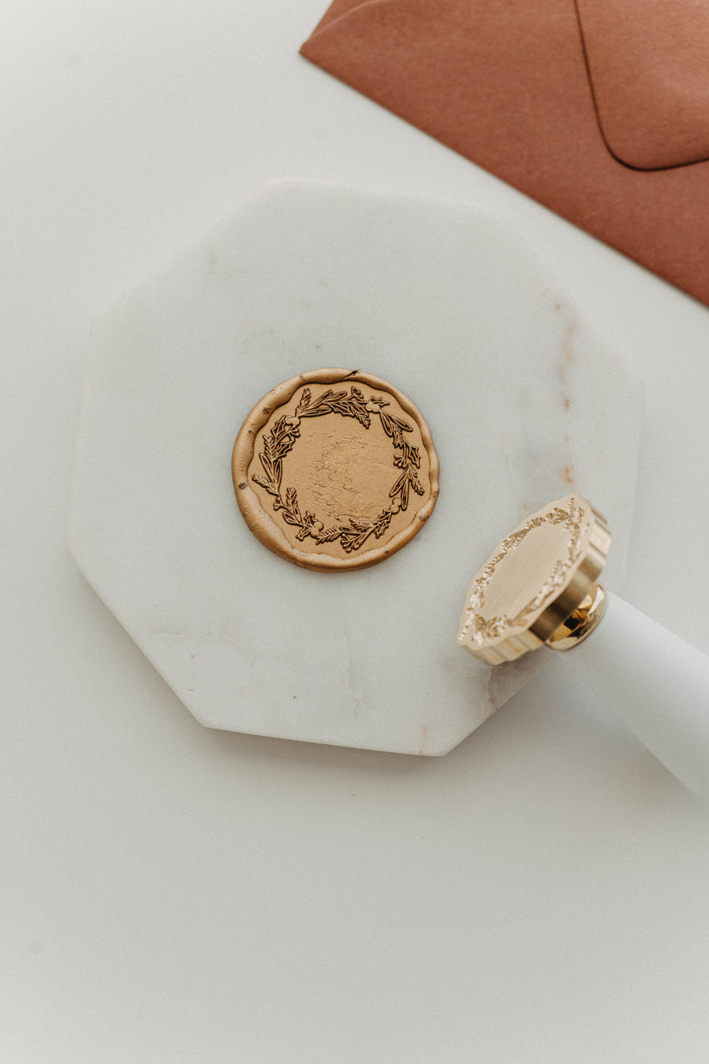 Metallic gold botanical wreath wax seal and white-handled brass wax stamp displayed on a white marble tile.