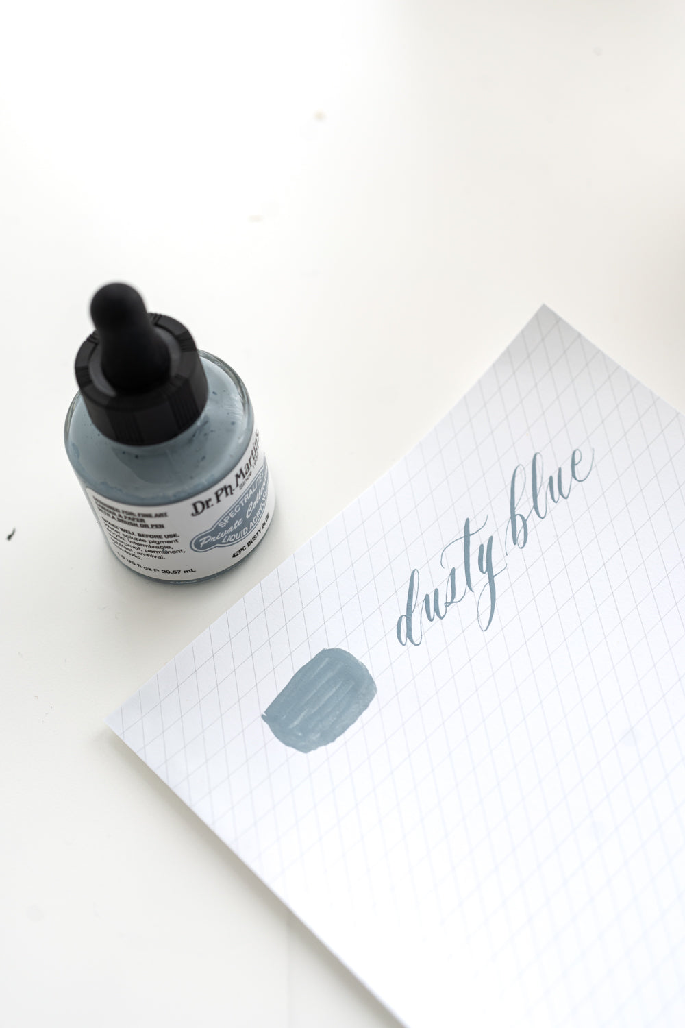 Dusty blue calligraphy ink swatch and bottle with lettering sample on grid paper for hand lettering