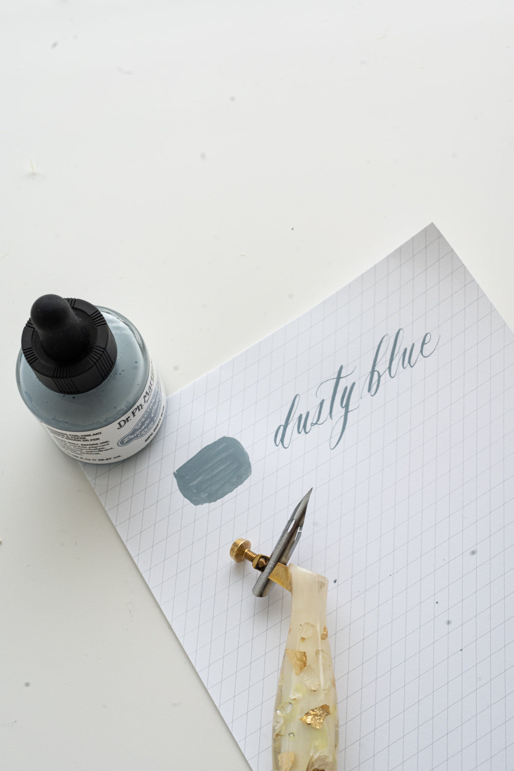Dusty blue calligraphy ink swatch with dip pen and nib on grid paper for modern calligraphy practice