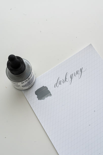 Dark gray calligraphy ink swatch on grid paper with Dr Ph Martin’s ink bottle, lettering practice setup