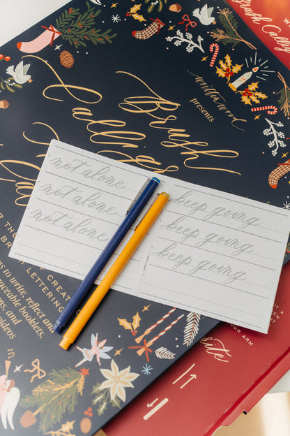Christmas calligraphy practice booklet and festive Advent Calendar Volume II with blue and yellow pens.