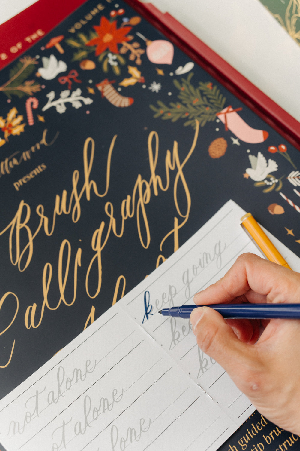 Hand practicing blue brush lettering in a guided Christmas calligraphy workbook beside a festive Advent calendar.
