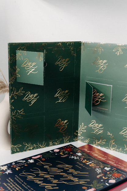 Open green Advent calendar with numbered doors and gold calligraphy, displayed beside a Christmas lettering booklet.