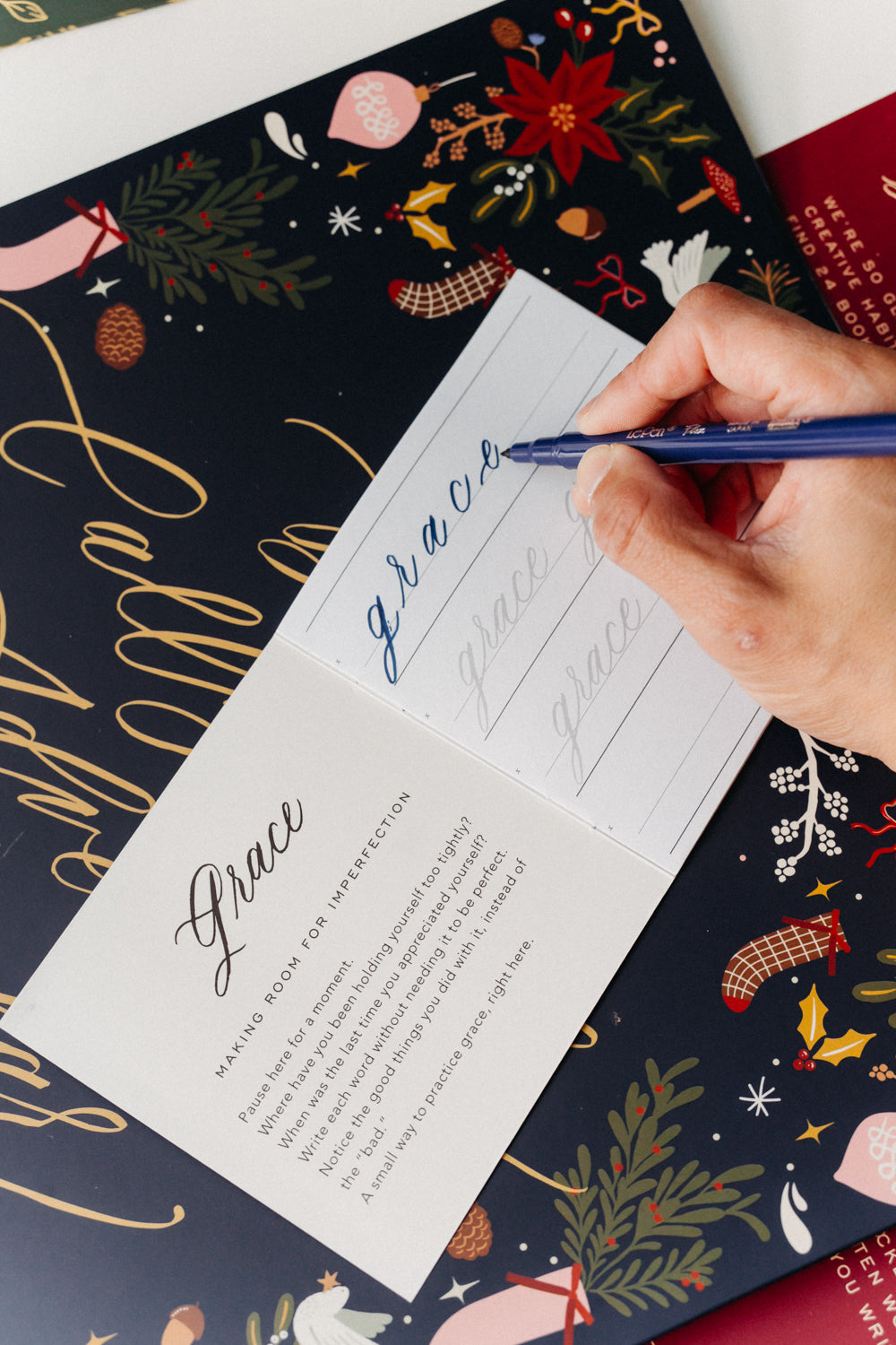 Hand using a blue brush pen to practice the word “grace” in a Christmas calligraphy Advent calendar workbook.