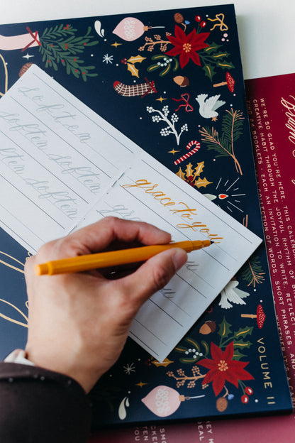 Hand practicing “grace to you” calligraphy in an Advent Calendar Volume II holiday lettering workbook.