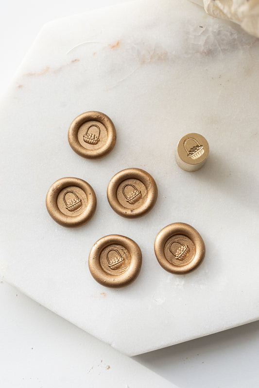 Close-up of gold sealing wax stamps with a 3d illustration of a basket of eggs made using a mini wax seal stamp on a marble background.